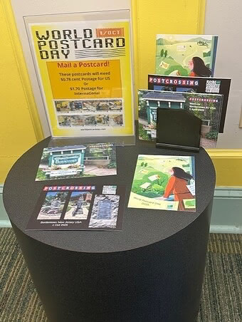 Display table with postcards and a sign promoting World Postcard Day on October 1. The sign includes postage information for sending postcards domestically and internationally