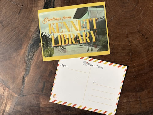 Two postcards on a wooden table, one reading Greetings from Kennett Library with a photo of the library building, and one showing a blank address side